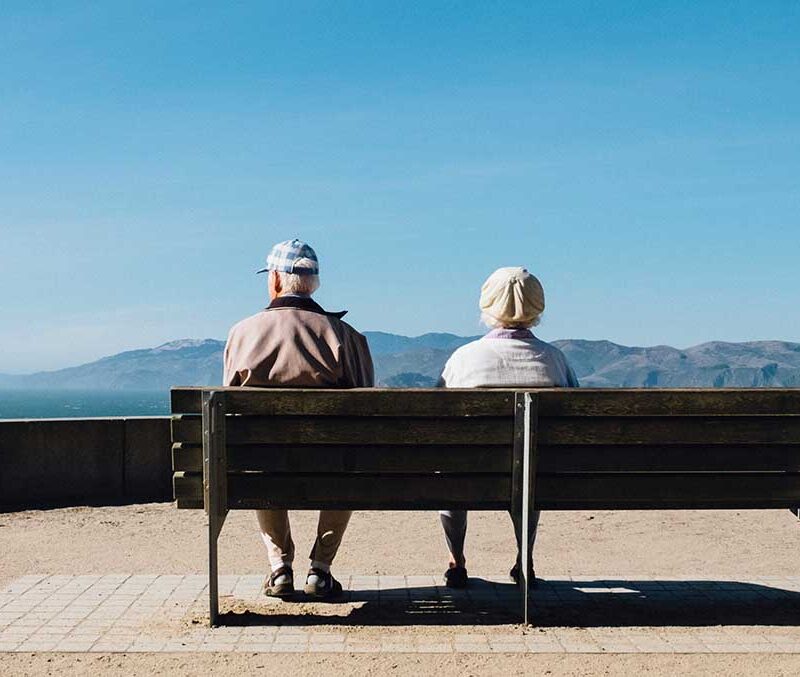 An elderly couple sitting on a bench looking atg mountains.